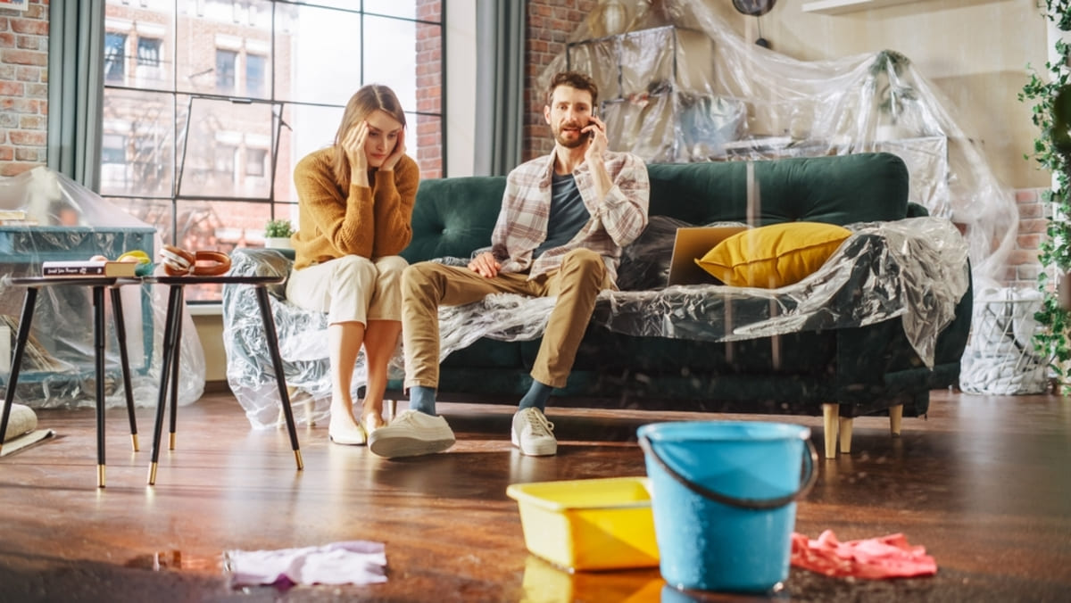 A couple on the couch calling someone surrounded by water and buckets, companies that buy homes for cash concept