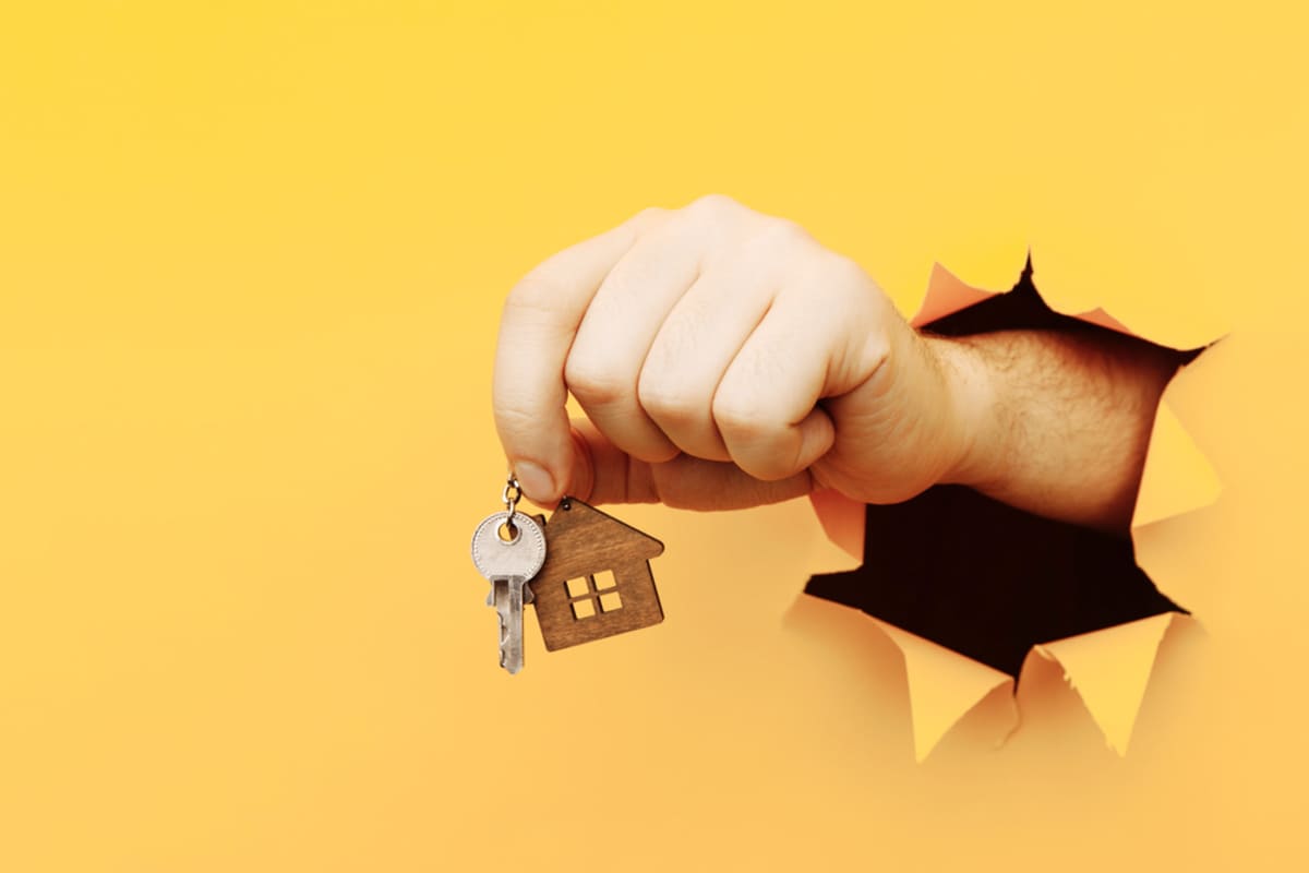 A hand coming through a yellow background holding house keys - buy my house for cash concept
